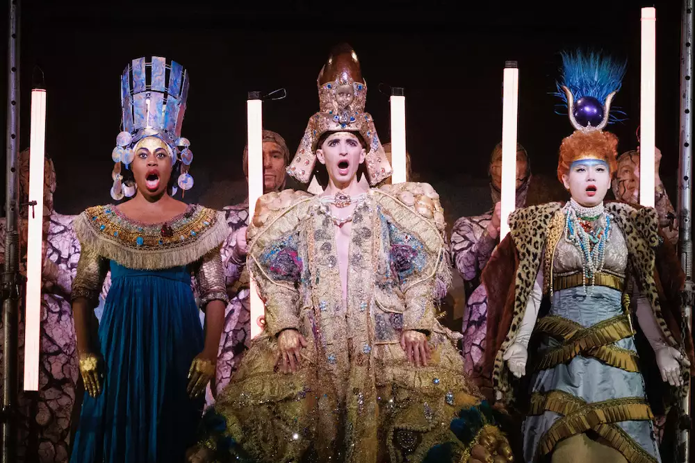 Three people on stage singing wearing elaborate jewelled dresses and headdresses