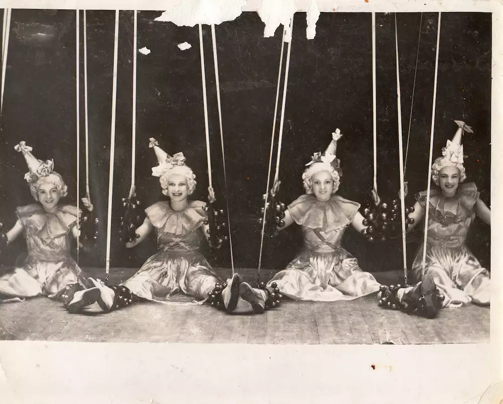 Four performers in matching costumes sit in a row, holding ropes like marionettes during a stage act.