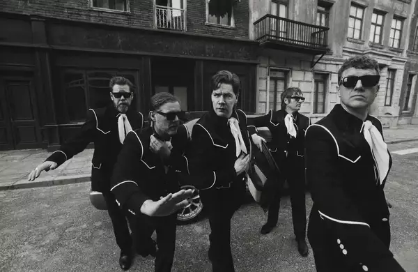 Black and white photograph of the five members of The Hives wearing matching suits and sunglasses striking dynamic poses on a city street. There is a car in the background.
