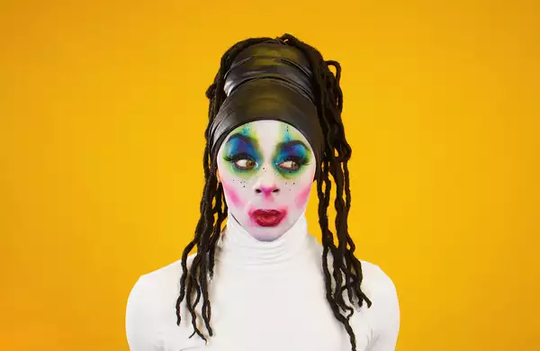 A person looking to the side wearing bright makeup and a white turtleneck top – on a bright orange background