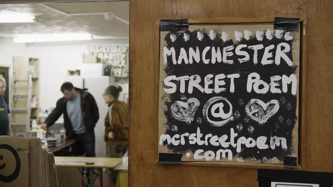 A sign saying Manchester Street Poem in the foreground with three people in the background working in a studio space
